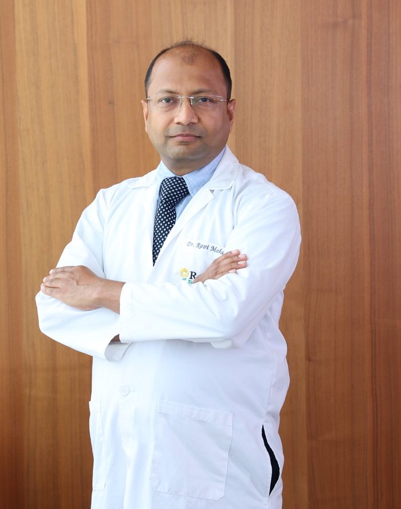 Dr. Ravi Mohanka in a white coat, arms folded, standing in front of a wooden wall